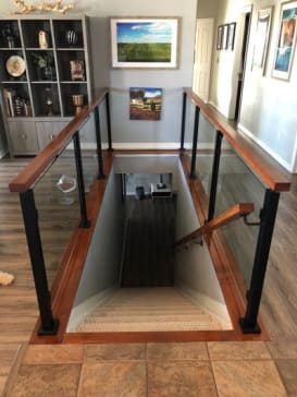 Black posts with glass panels and a wood cap rail enclosing a stairwell opening on a tiled floor.