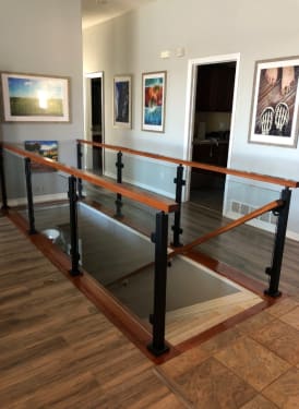 Black posts with glass panels and a stained wood top rail surround a stair opening in a hallway with framed art.