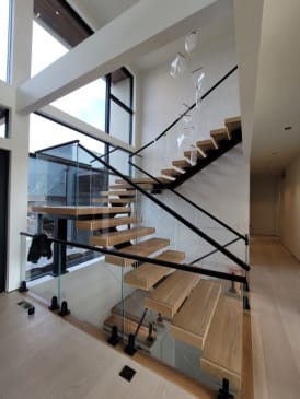 Light wood treads on a black mono stringer with glass railing climb a tall window-lit stairwell.