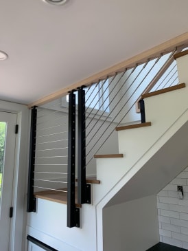 Black posts with stainless cable infill rising along a white stair with oak treads to a wood ceiling header.