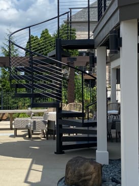 Black steel spiral stair with curved cable railing beside a white column and patio seating.