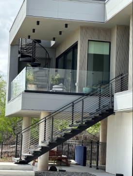 Elevated modern home with a black exterior stair with cable railing, glass balcony railing and spiral stair above.