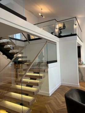 Light oak floating treads with glowing under-tread LEDs climb beside frameless glass with a wood cap rail to a glass-railed loft.