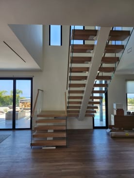 White oak floating stair with glass railings rising through a bright great room with ocean views