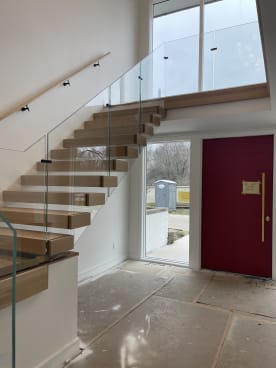 White oak cantilever treads with floor-to-ceiling frameless glass and an oak wall handrail beside a red front door