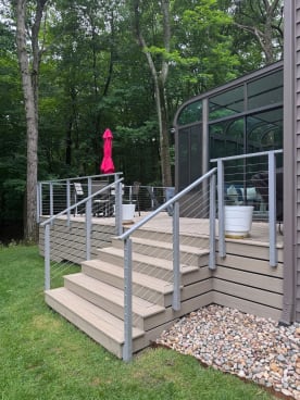 Wide composite deck stairs with gray-post cable railing beside a sunroom in a wooded yard.