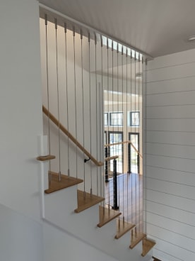 Vertical stainless cables run floor to ceiling beside an oak-tread stair with a wall-mounted wood handrail.