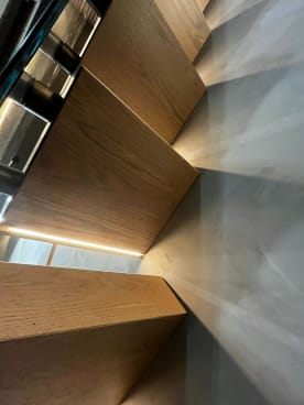 Underside view of white oak cantilever treads with LED strips casting light across a plaster wall.