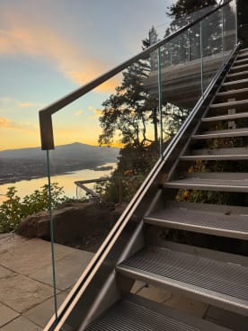 Frameless glass railing with a bronze handrail along an exterior steel stair overlooking a river valley at sunset.