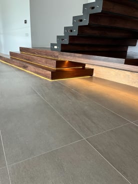 Stacked walnut plinth steps with warm LED under-glow at the foot of a cantilevered walnut stair on tile floor.