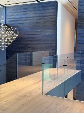 Frameless glass panels line an oak bridge beside a dark-planked wall and sparkling chandelier