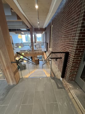 Frameless glass with black handrails around a stair opening beside exposed brick and timber columns