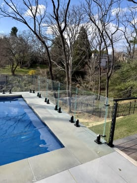 Frameless glass on black talon spigots fences a pool deck, meeting black cable railing among mature trees