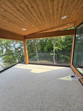 Frameless base rail glass wraps a covered cedar-ceiling porch overlooking a wooded lake