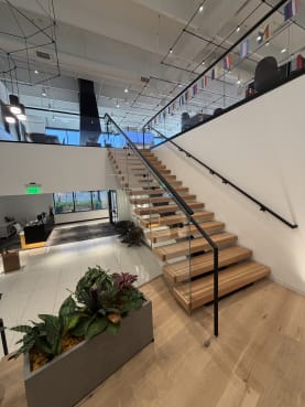 Wide wood floating treads with frameless glass and black handrails rising through a bright office lobby