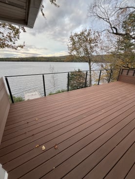 Black cable railing wraps a composite deck above an autumn lake shore with a dock below