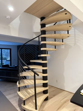 A light maple spiral stair around a black center column with a sweeping black cable railing in a bright room.