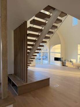 A FLIGHT Mono stair with oak treads and frameless glass railing rises beside a wood slat partition in a tall arched room.