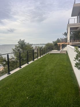 Black cable railing runs along a cliff-top lawn above the water beside a modern home.