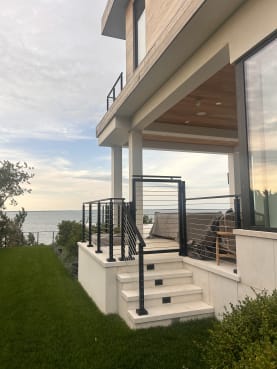 Black cable railing wraps a stone porch and steps of a modern home overlooking the water.