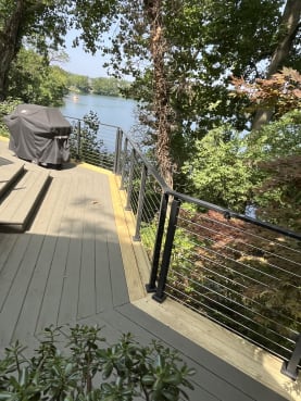 Black cable railing wraps a gray deck corner overlooking a lake, with a covered grill nearby.