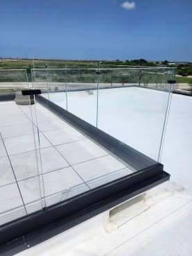Frameless glass panels in a dark base-rail channel around a white rooftop deck with open coastal views.