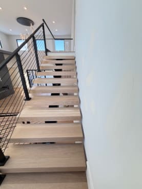 White oak floating treads rise along a white wall beside a black cable railing.