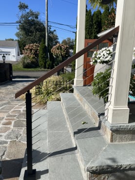 Cable railing with a wood handrail guards bluestone porch steps beside white columns and flowers.