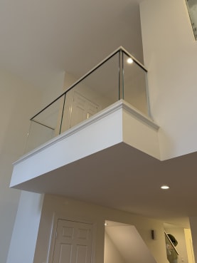 A frameless glass railing with slim cap tops a white interior balcony above a doorway.