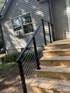 A black cable stair railing follows cedar entry steps up to a landing beside gray siding.