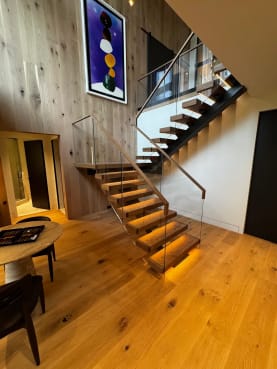 Thick oak treads with warm under-tread LED lighting float on black stringers beside frameless glass with a wood handrail in a double-height wood-paneled room.