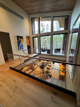 A frameless glass guard with wood cap overlooks a double-height living room with floor-to-ceiling windows.