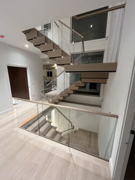 A U-shaped stack stair of thick white oak treads with LED step lighting rises between frameless glass guards with a wood handrail.