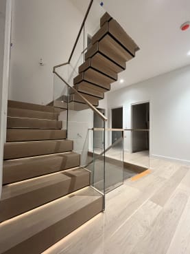 A U-shaped stack stair of thick white oak treads with LED step lighting rises between frameless glass guards with a wood handrail.