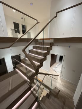 A U-shaped stack stair of thick white oak treads with LED step lighting rises between frameless glass guards with a wood handrail.