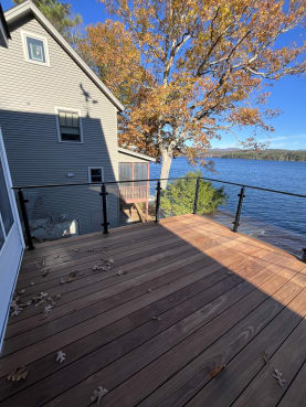 Black posts hold glass panels along an ipe deck overlooking a lake with autumn trees.