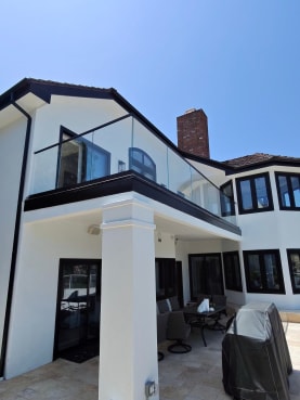 A frameless glass guard tops a black-fascia balcony on a white stucco home above a stone patio.