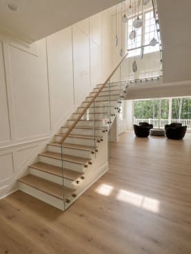Frameless glass panels with brass standoff pins and an oak handrail line a white-oak stair in a paneled white entry.