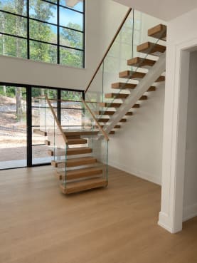 White oak cantilevered treads float off a white wall beside a frameless glass guard with oak handrail in front of tall black steel windows.
