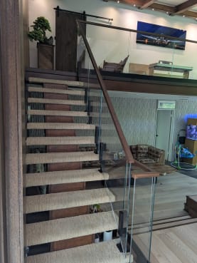 Frameless glass panels with a dark wood handrail flank an open-riser stair with woven tread runners.