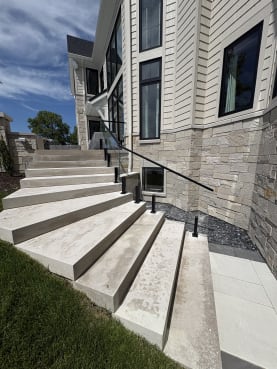 Black talon clamps hold frameless glass with a black top rail along sweeping limestone exterior steps beside a stone-clad home.