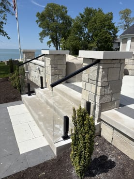 Frameless glass in black talon clamps with a black handrail flanks limestone steps between stone pillars near a lake.
