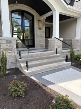 Black talon-clamped glass rails with black handrails flank limestone steps leading to an arched front door of a stone home.