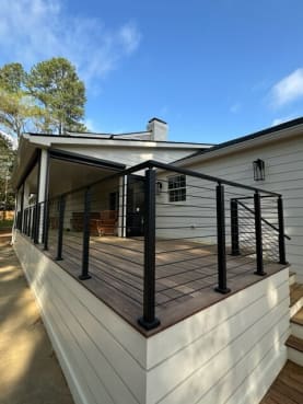 Black posts with horizontal rods wrap the corner of a raised wood porch on a white-sided home.
