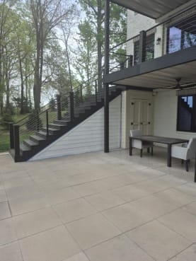 Black cable railing follows a dark composite exterior stair up to a balcony beside a covered patio.