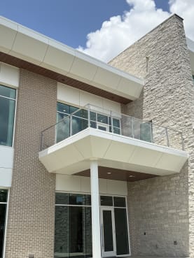 Post-to-post glass railing on a white cantilevered balcony above the entrance of a brick and stone office building.