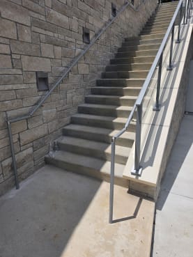 Post-to-post glass and metal handrails along an exterior concrete stair beside a stone wall.