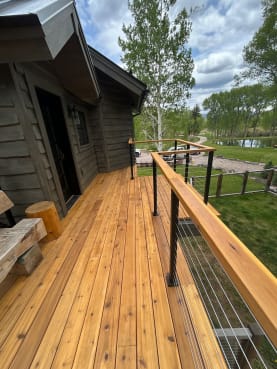 Cedar deck with cedar top rail and black-post cable railing beside a dark wood cabin and green pasture.