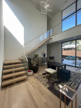 White oak floating treads with glass railing climb to a bridge in a sunlit double-height living room.