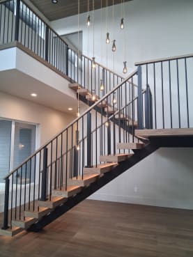 Wood treads on a black mono stringer with black vertical baluster railing climb to a balcony under drop pendants.